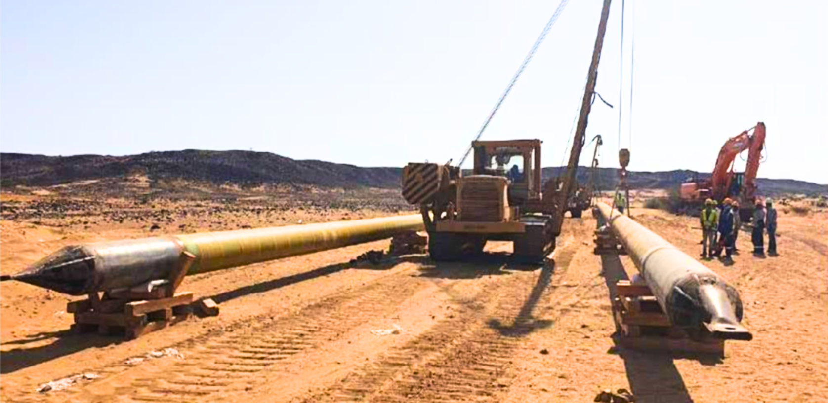 Large cylindrical horizontal directional drilling pipes being transported on a dirt road with construction equipment.